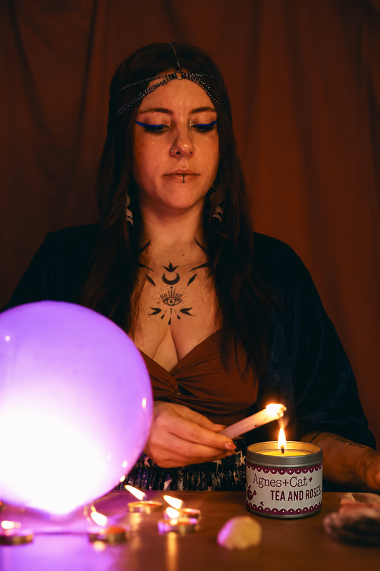Woman with tattoos holding a lit candle in front of a crystal ball and other objects on a table.