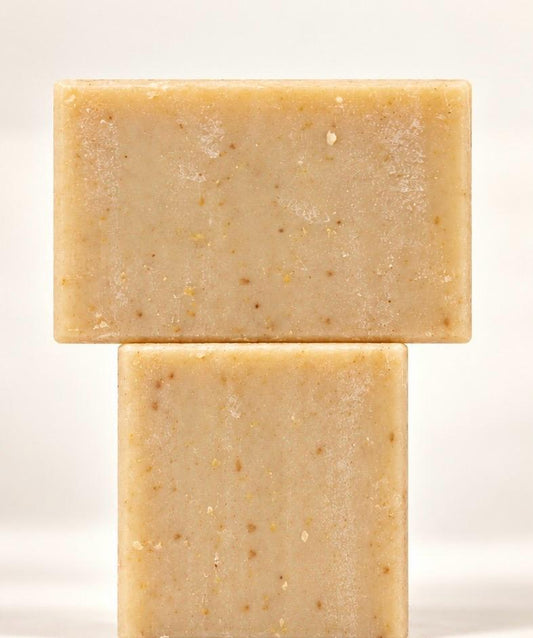 Two honey and oatmeal soap bars stacked in a T shape on a cream background, with a soft reflection beneath.