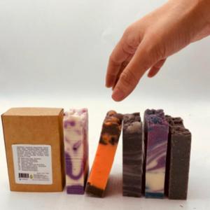 Hand reaching for a stack of colorful soap bars with a brown box on a white background
