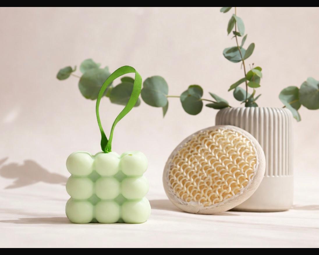 Light green massage soap bar with textured bubble design and hanging strap in a minimal spa setting with eucalyptus leaves and natural body brush