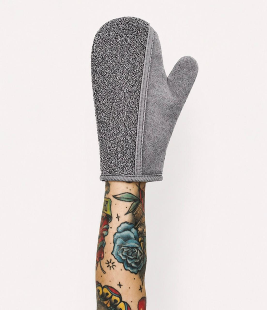 Gray exfoliating bath mitt on a tattooed arm, showing soft fabric texture and spa body care design.
