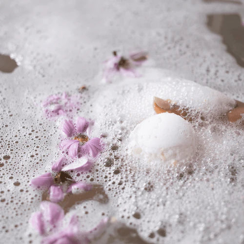 Lemon bath bomb dissolving in foamy bath water with delicate flower petals, creating a relaxing spa atmosphere and fresh citrus bathing experience.