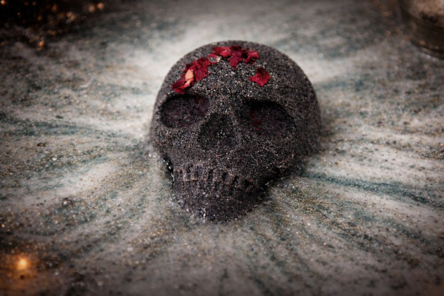 Skull bath bomb with red flowers on a textured surface dissolving in water, creating dark swirling foam – Halloween bath bomb by Lumaysa