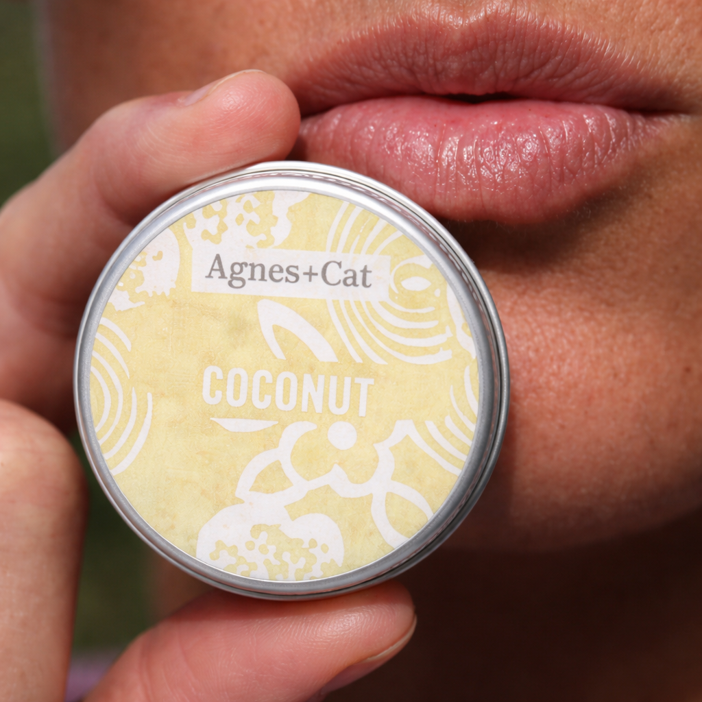 Close-up of Agnes & Cat Coconut lip balm tin held near lips, showcasing natural moisturizing lip care with a soft glossy finish.
