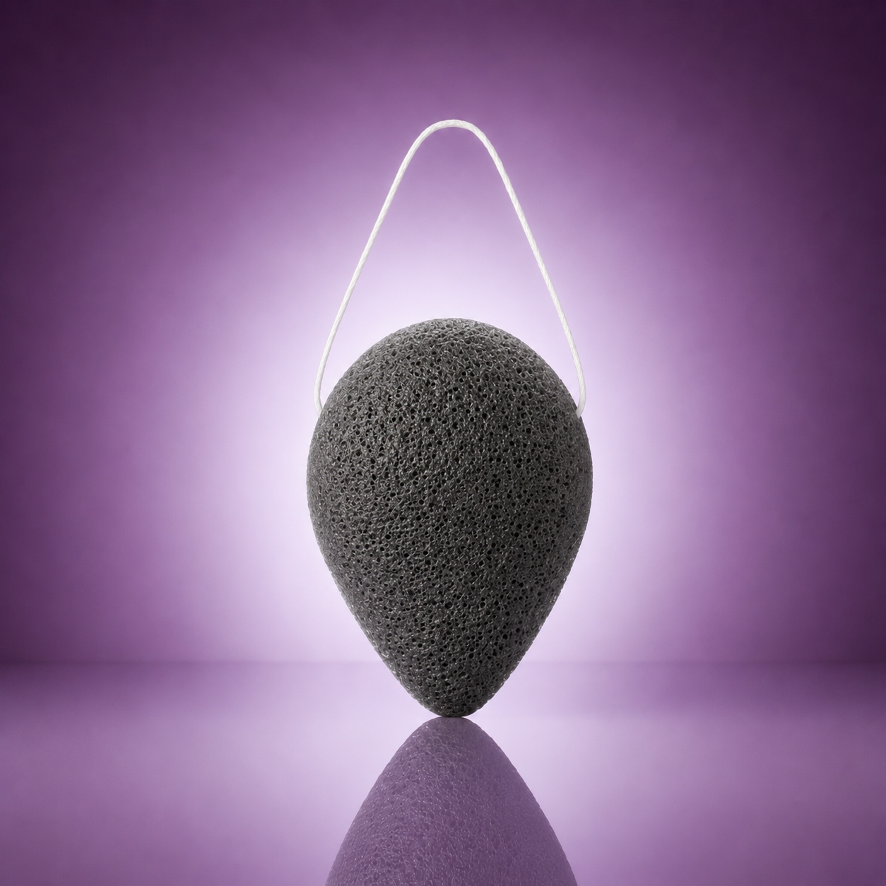 Purifying bamboo charcoal konjac sponge with a white hanging string on a soft purple background.