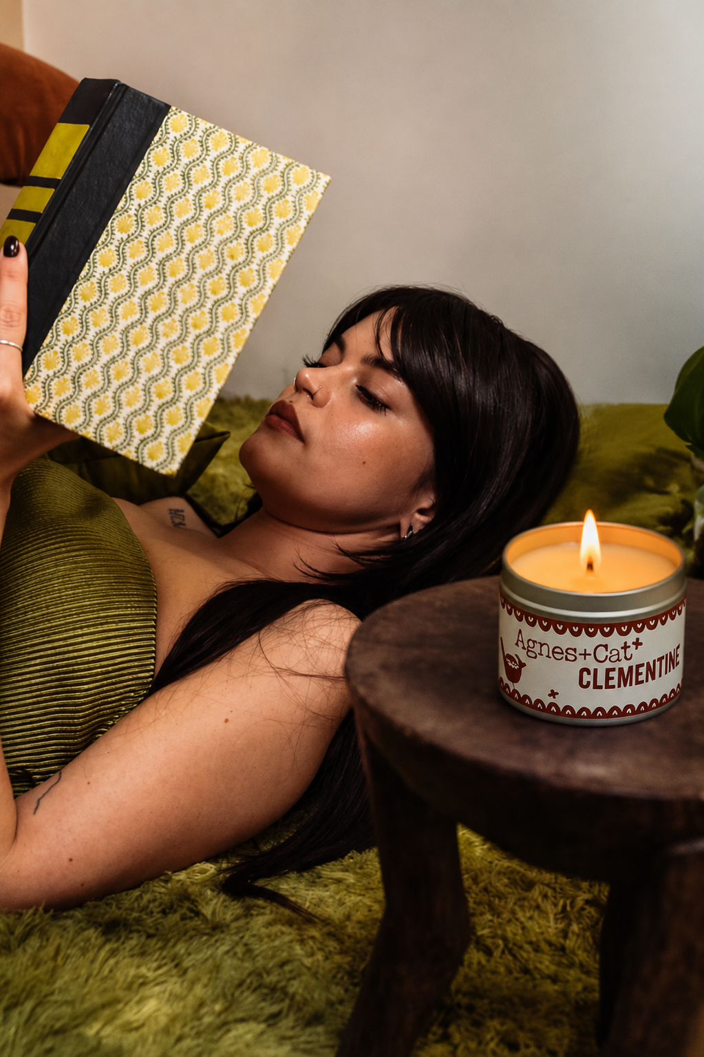 Lit Agnes + Cat Clementine soy candle on a wooden table beside a woman reading in a cozy relaxing home setting.