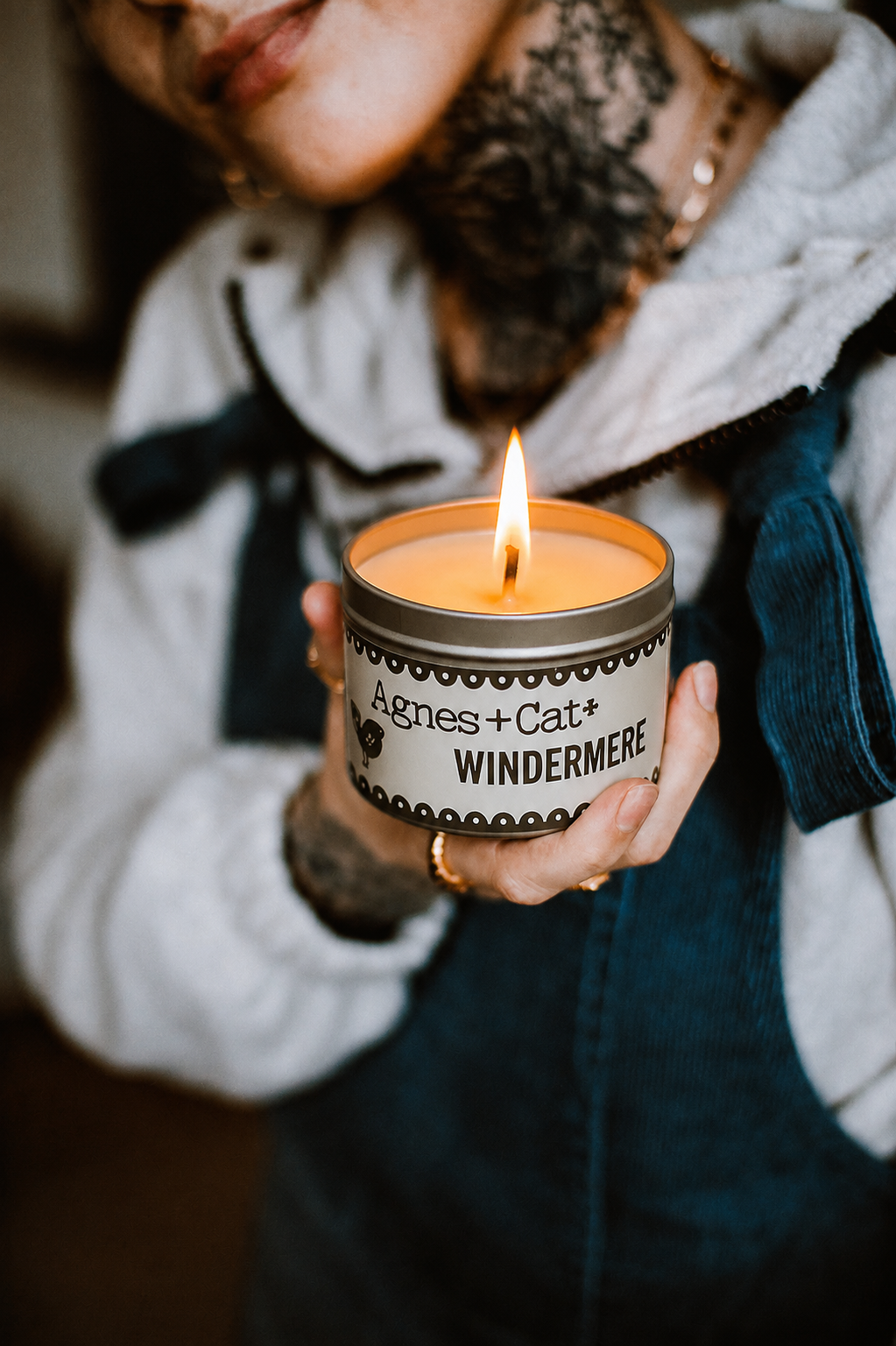 Person holding a lit candle in a container labeled 'Agnes + Cat Windermere'.