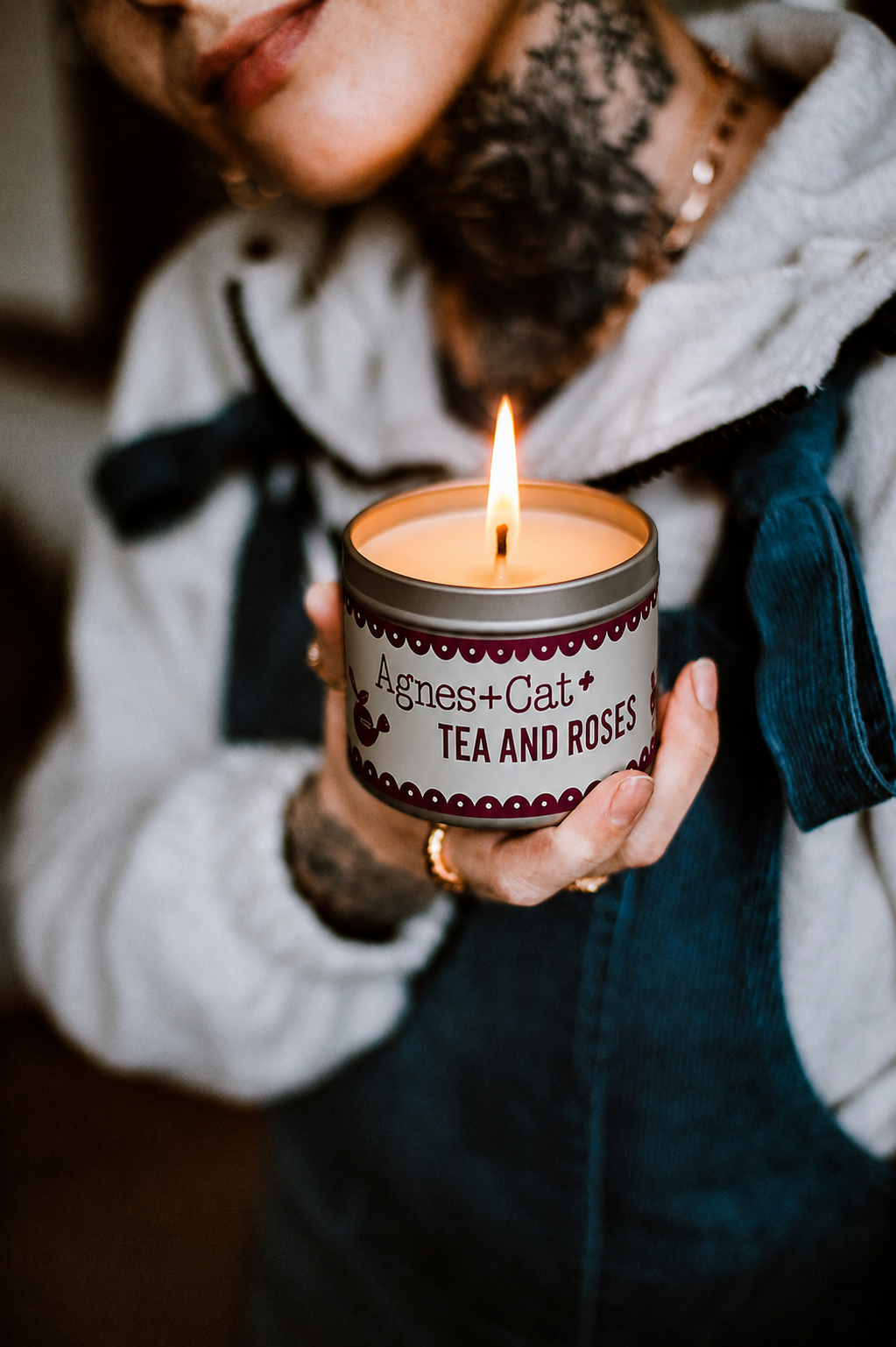 Person holding a lit candle in a tin labeled 'Agnes + Cat Tea & Roses' against a blurred background.