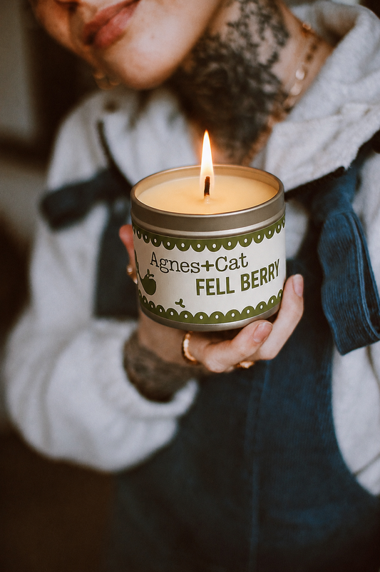 Person holding a lit candle in a tin labeled 'Agnes + Cat Fell Berry'.