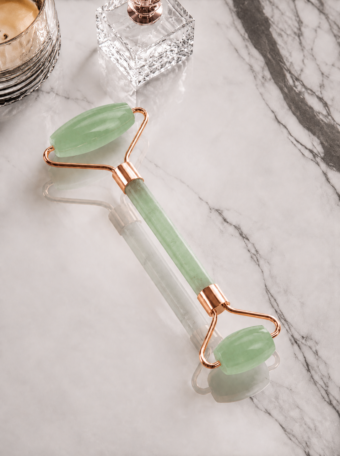 Jade facial roller on a marble surface with a candle and perfume bottle in the background.