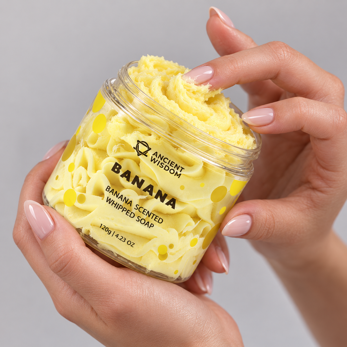 Hands holding an open jar of Banana Scented Whipped Soap 120g and touching its creamy yellow texture