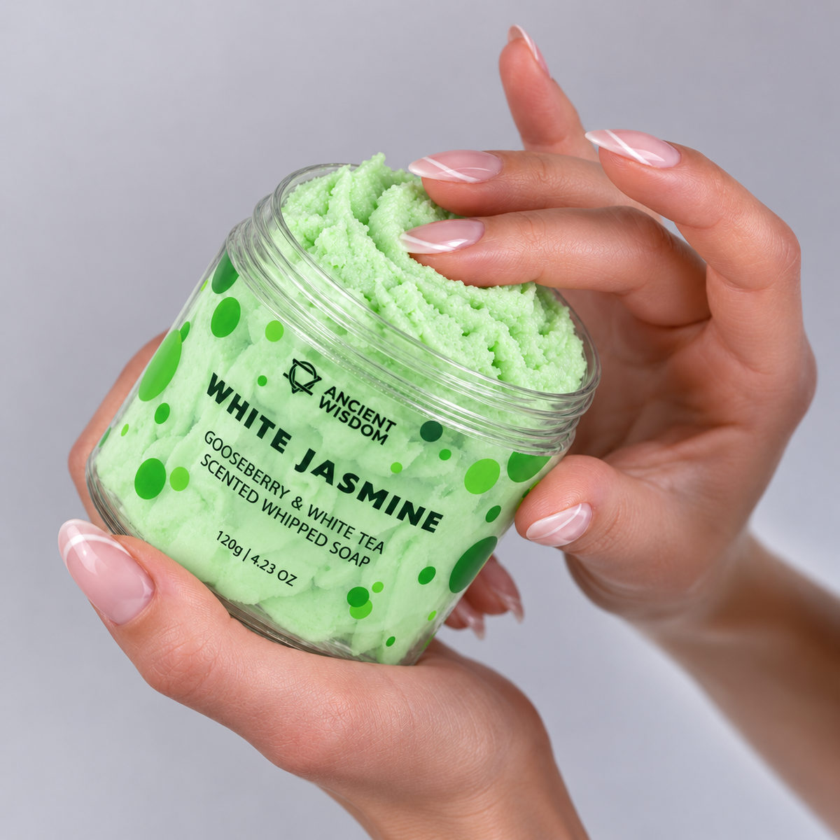 Hands holding an open jar of pale green White Jasmine Whipped Soap and gently touching its creamy texture.