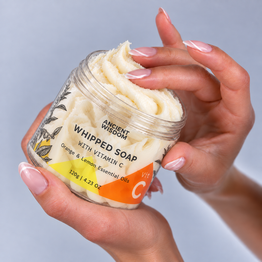 Woman holding an open jar of Ancient Wisdom Whipped Soap with Vitamin C and orange and lemon essential oils, featuring a creamy ivory texture.