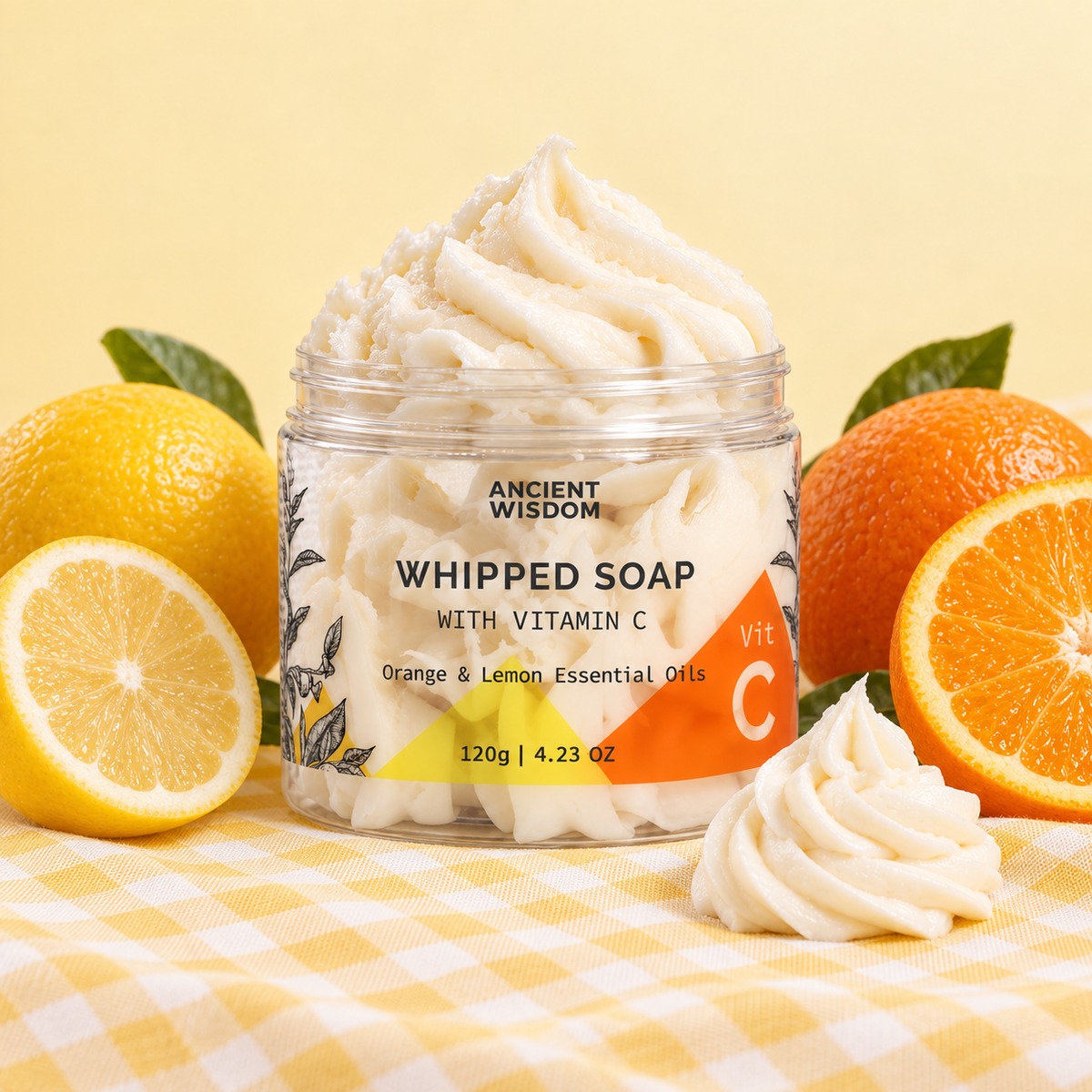 Open jar of Ancient Wisdom Whipped Soap with Vitamin C, displayed with fresh oranges, lemons and a creamy soap swirl on yellow gingham fabric.