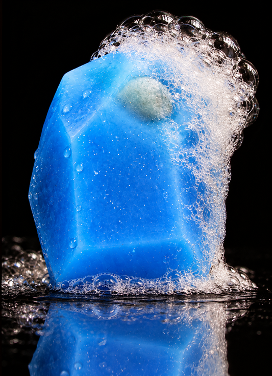 Blue crystal-shaped soap with an embedded gemstone, water droplets and natural lather cascading down one side on a black reflective surface.