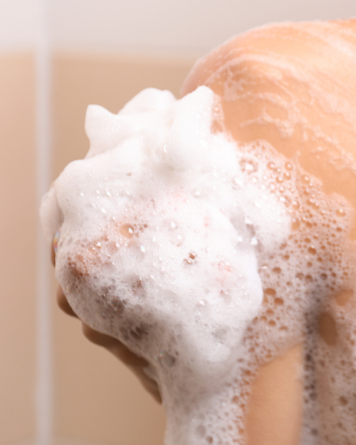 Hand holding a ramie shower puff covered in rich soap lather against wet skin.