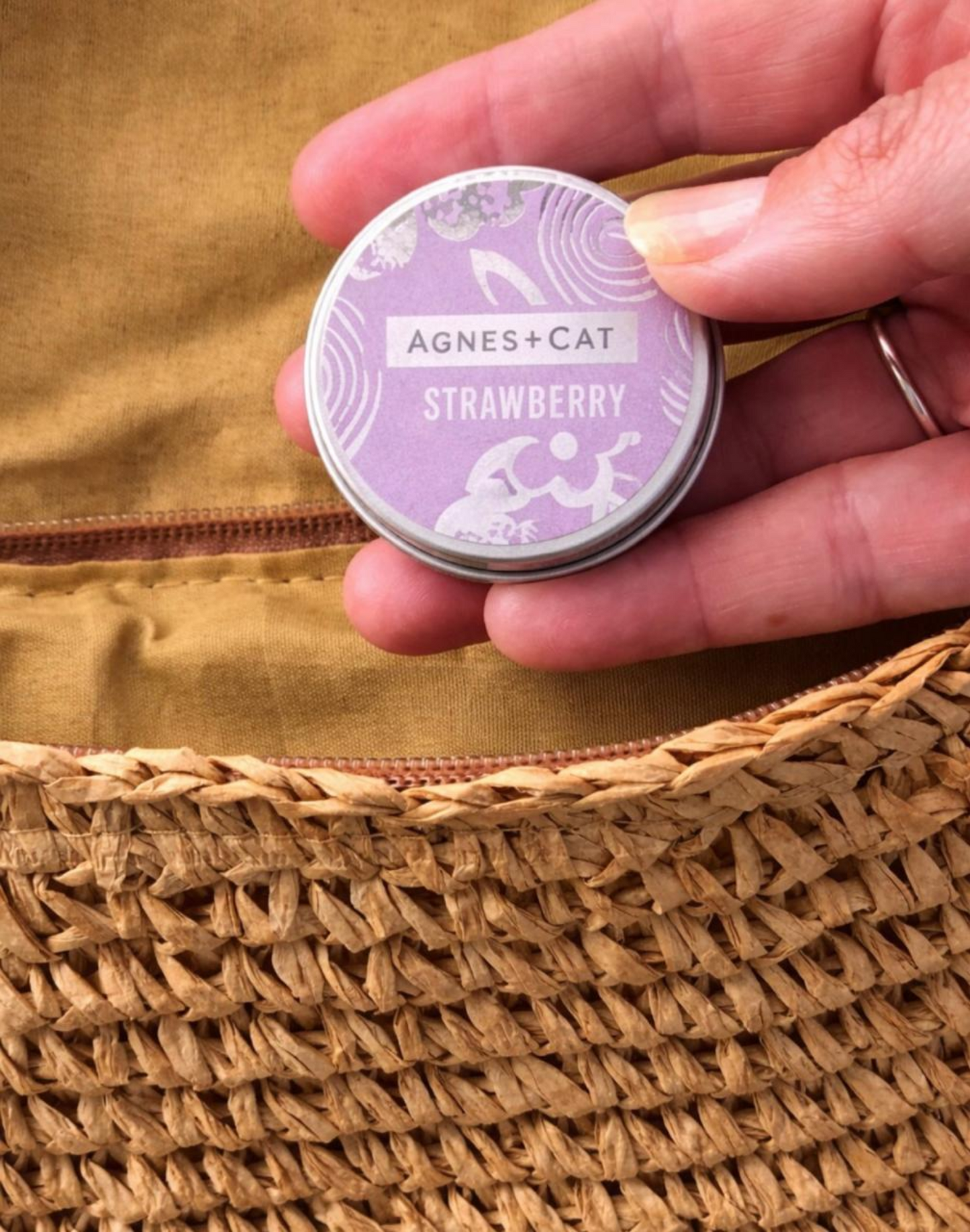 Close-up of a hand holding Agnes & Cat Strawberry lip balm tin with natural skincare packaging over a woven straw bag in warm sunlight.