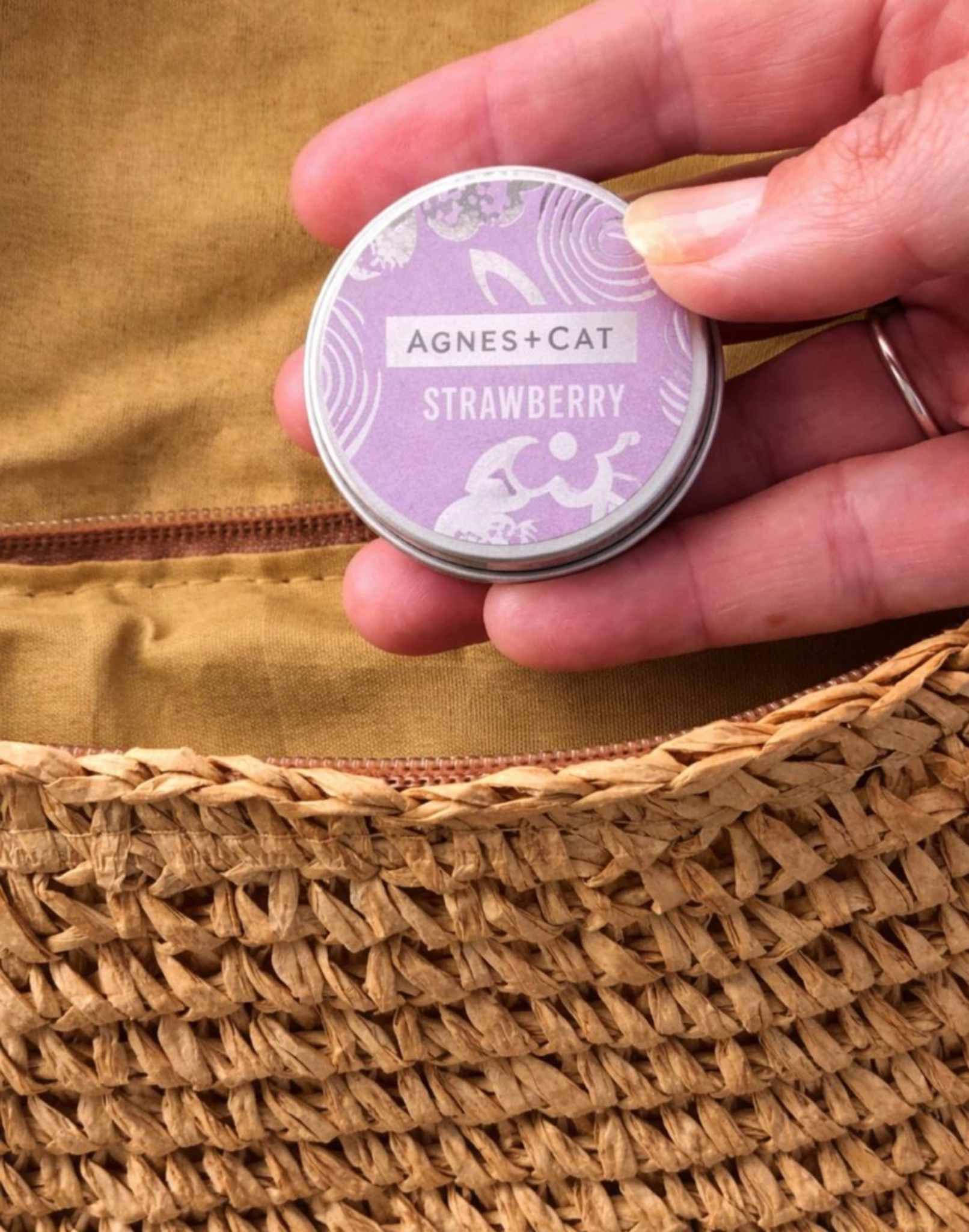 Close-up of a hand holding Agnes & Cat Strawberry lip balm tin with natural skincare packaging over a woven straw bag in warm sunlight.